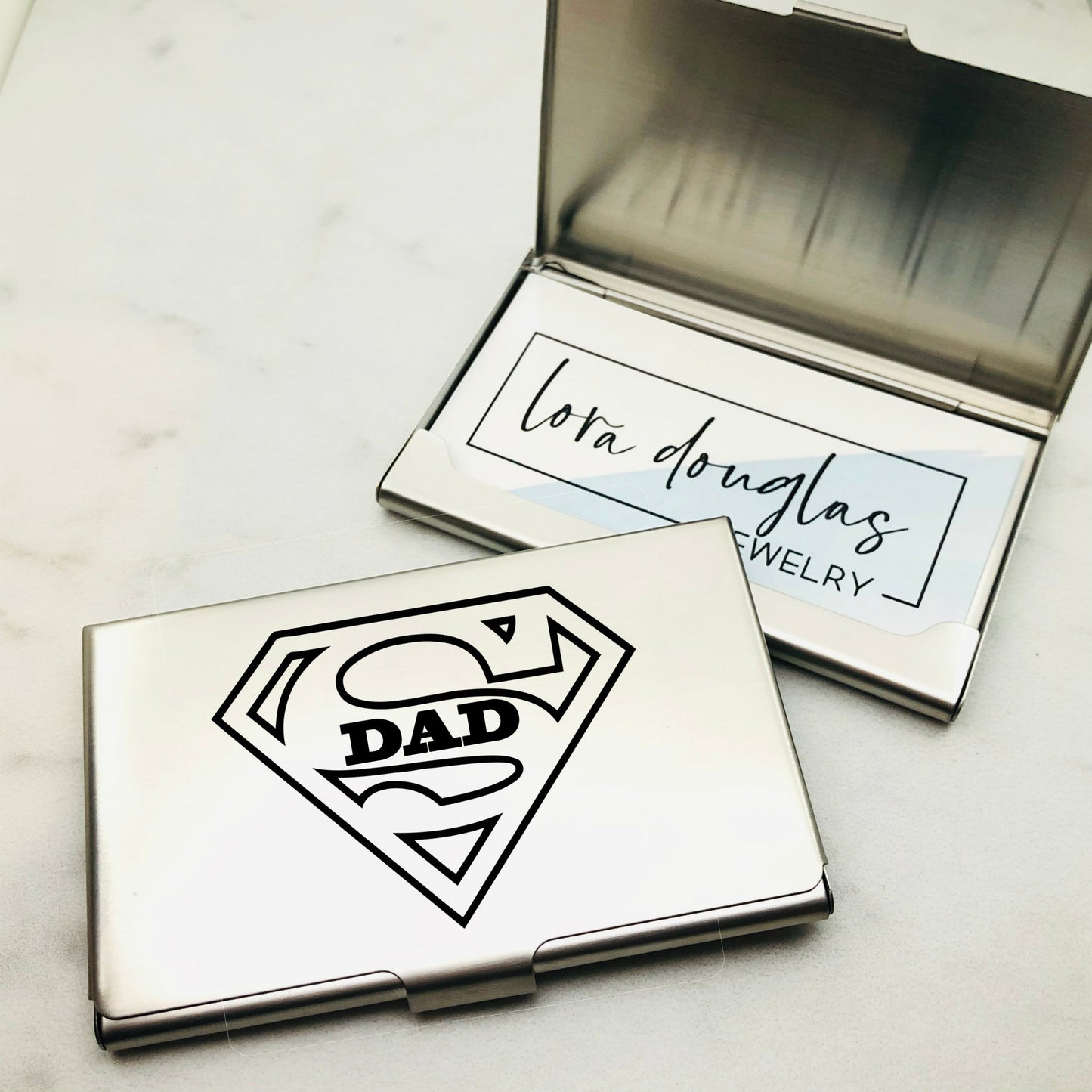 A stainless steel business card holder with an engraved design that reads DAD along with a graphic of a superhero shield.