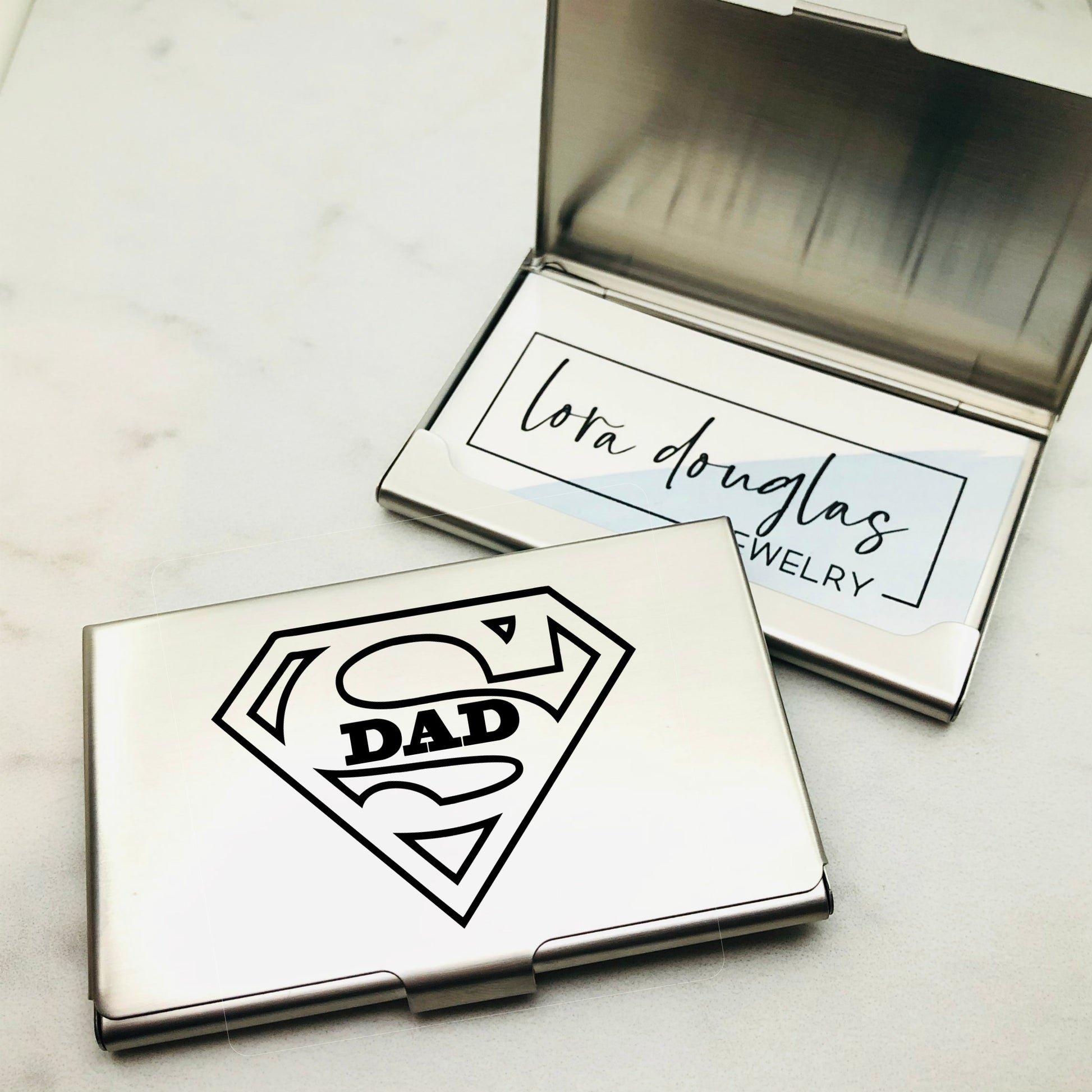 A stainless steel business card holder with an engraved design that reads DAD along with a graphic of a superhero shield.
