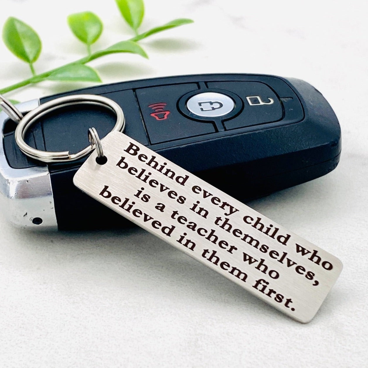 A stainless steel keychain with an engraved message 'BEHIND EVERY CHILD IS A TEACHER WHO BELIEVED IN THEM FIRST' placed on a car key. The keychain has a silver color and a standard key ring attached.