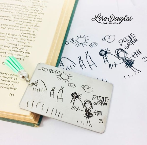 A custom stainless steel bookmark with a laser engraved child's drawing, presented next to its navy blue velvet drawstring pouch.