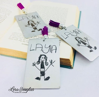 A custom stainless steel bookmark with a laser engraved child's drawing, presented next to its navy blue velvet drawstring pouch.